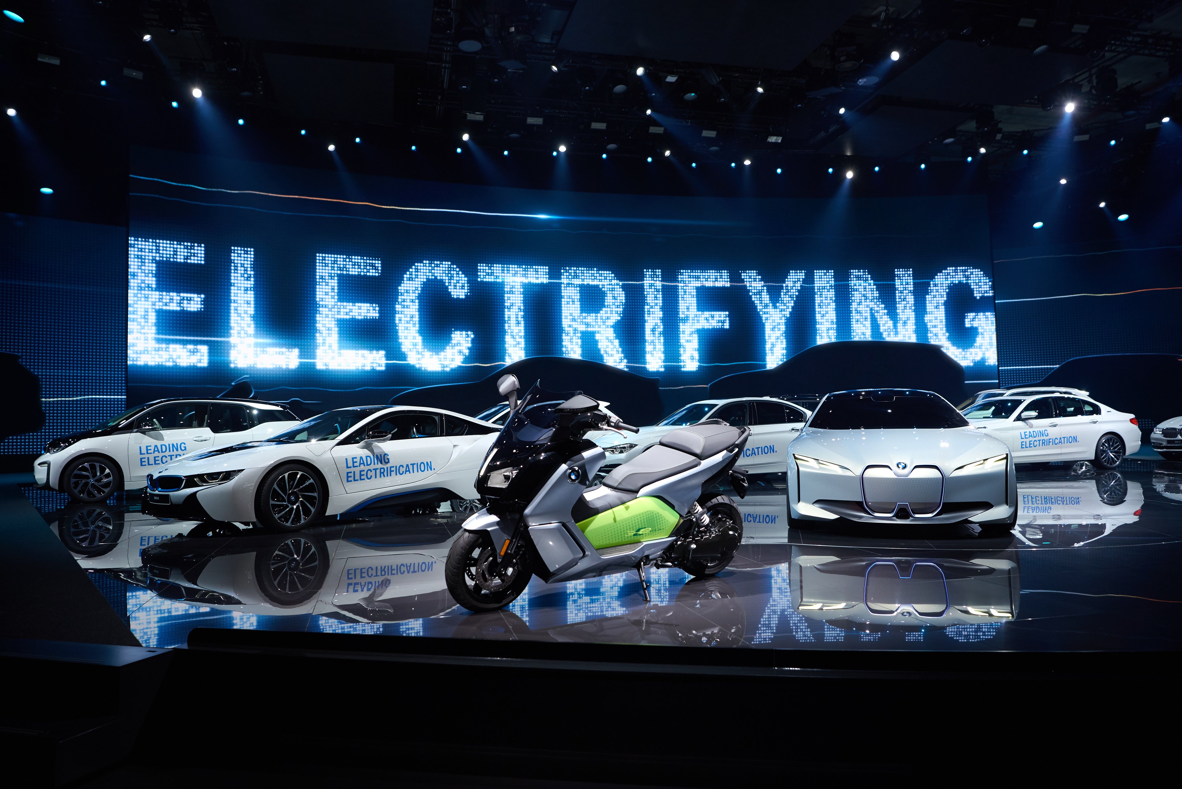 The i Vision Next is shown parked on a show stand, with other BMWs -- including an electric motorcycle -- parked nearby. The word "electrifying" appears on a big screen in the background.