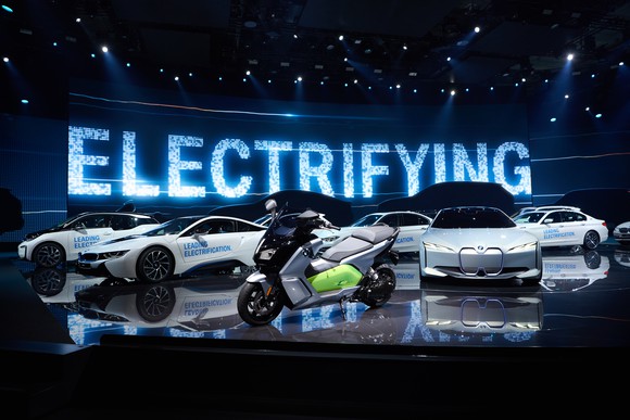 The i Vision Next is shown parked on a show stand, with other BMWs -- including an electric motorcycle -- parked nearby. The word "electrifying" appears on a big screen in the background.