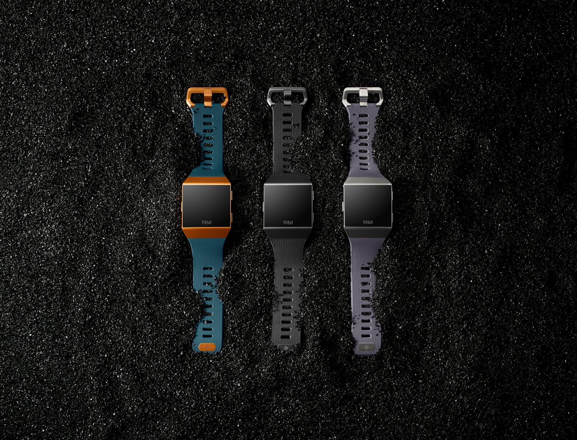 Three Fitbit Ionic smartwatches with different bands, laid out on a bed of a gravel-like substance.