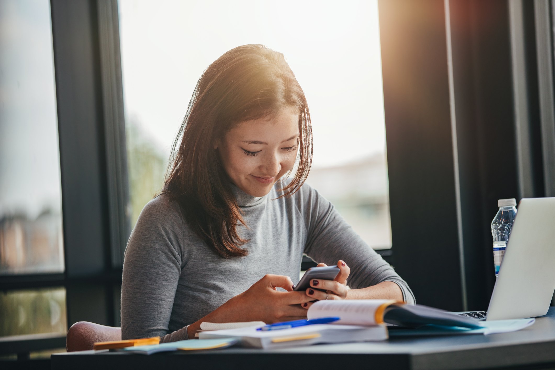 A woman sitting at a table using a smartphone. There are papers on the table.
