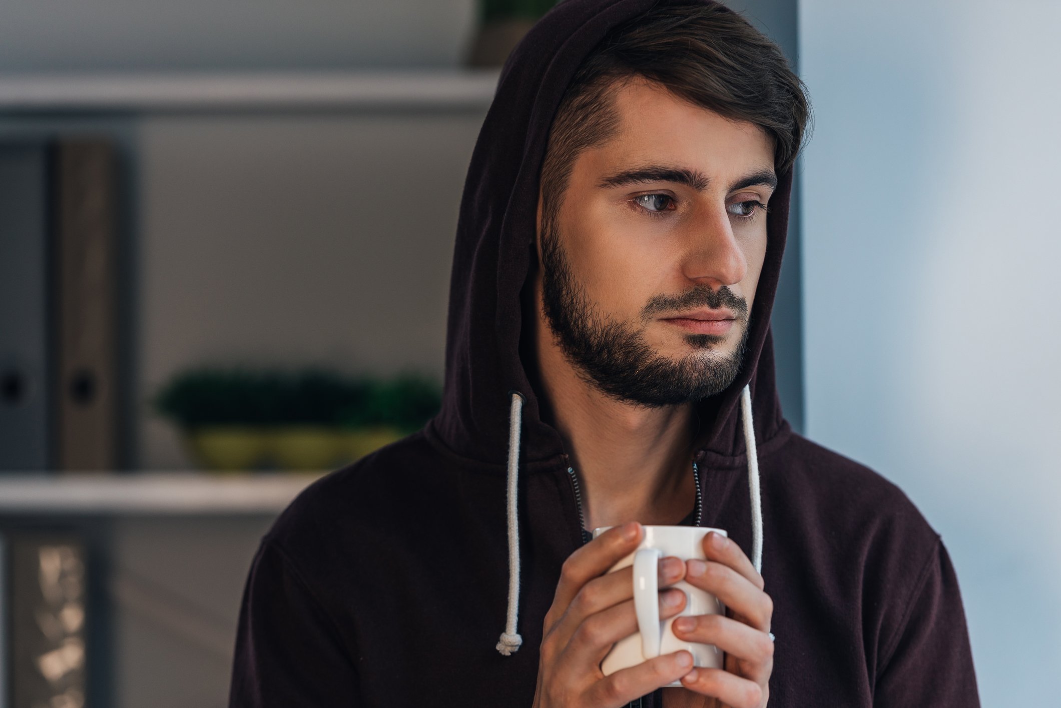 Serious younger man wearing a hoodie and clutching a coffee mug.