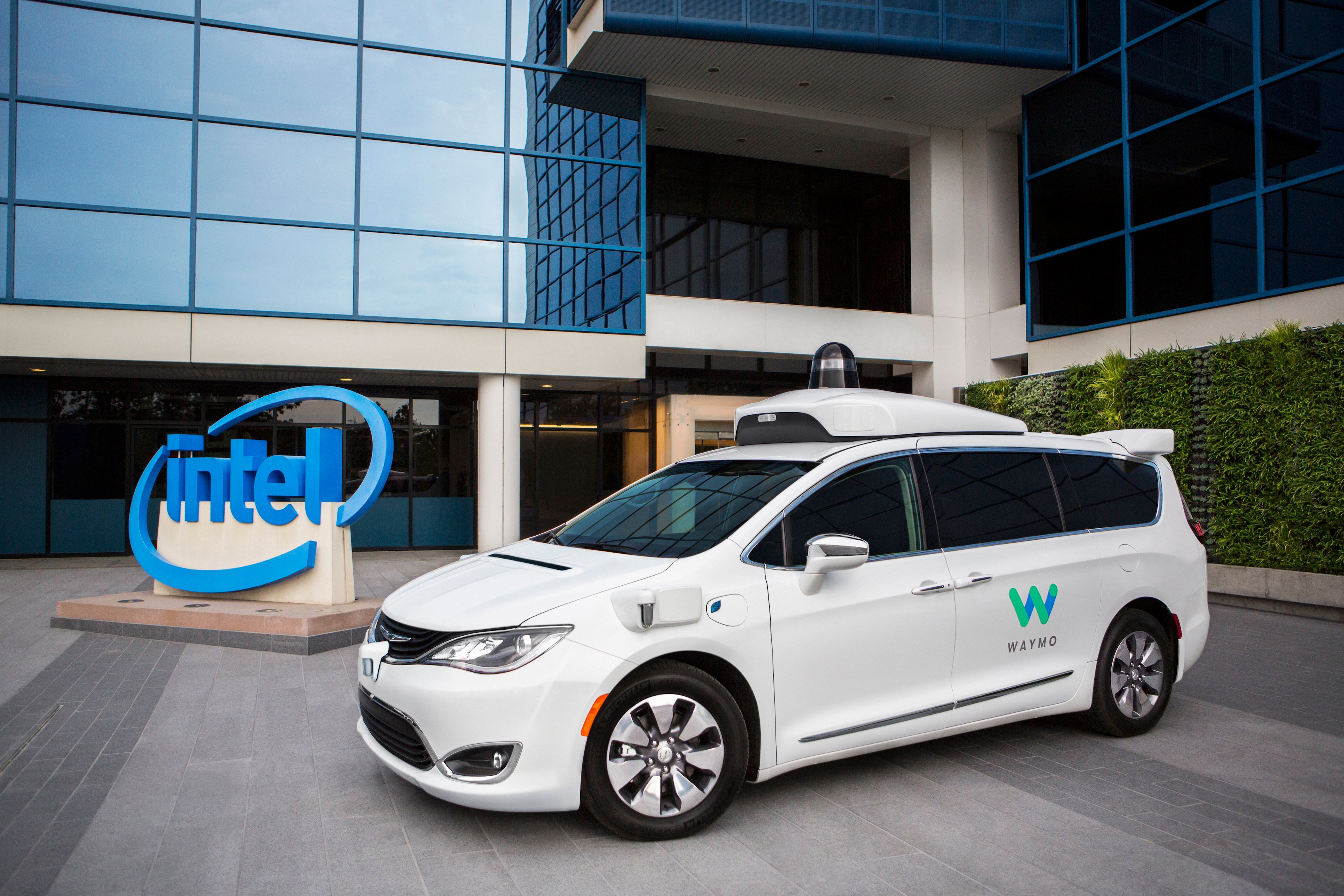 A white Chrysler Pacifica Hybrid minivan with Waymo logos and visible self-driving sensor hardware is parked in front of an Intel building.