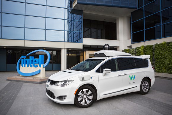 A white Chrysler Pacifica Hybrid minivan with Waymo logos and visible self-driving sensor hardware is parked in front of an Intel building.