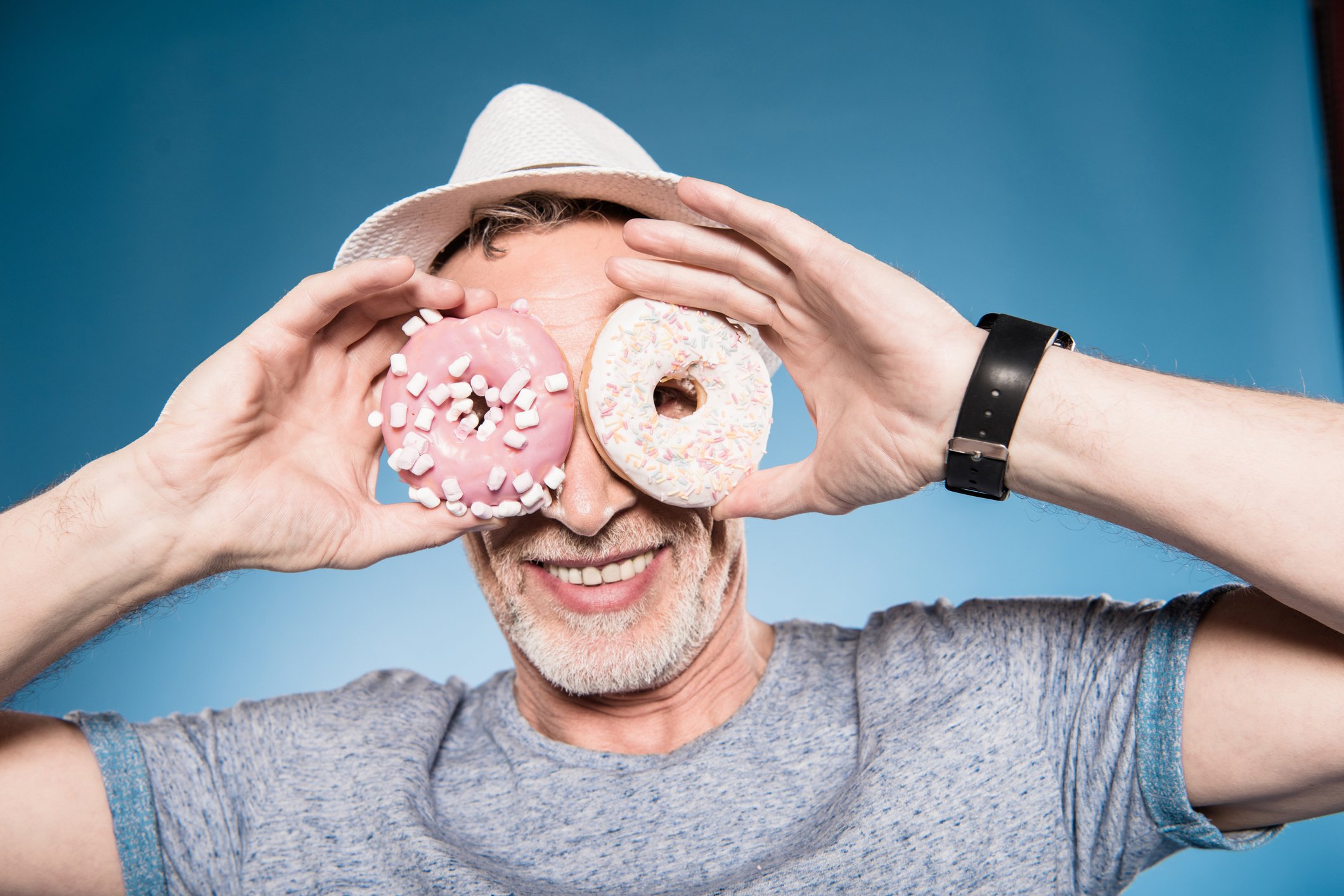 A senior man looking through the holes of two donuts he's holding in his hands.