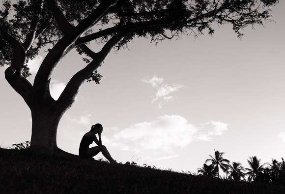 The silhouette of a woman with her hands in her face, on a hill, by a tree.