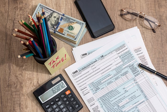 Tax forms, calculator, pencils, and stack of money on a table.