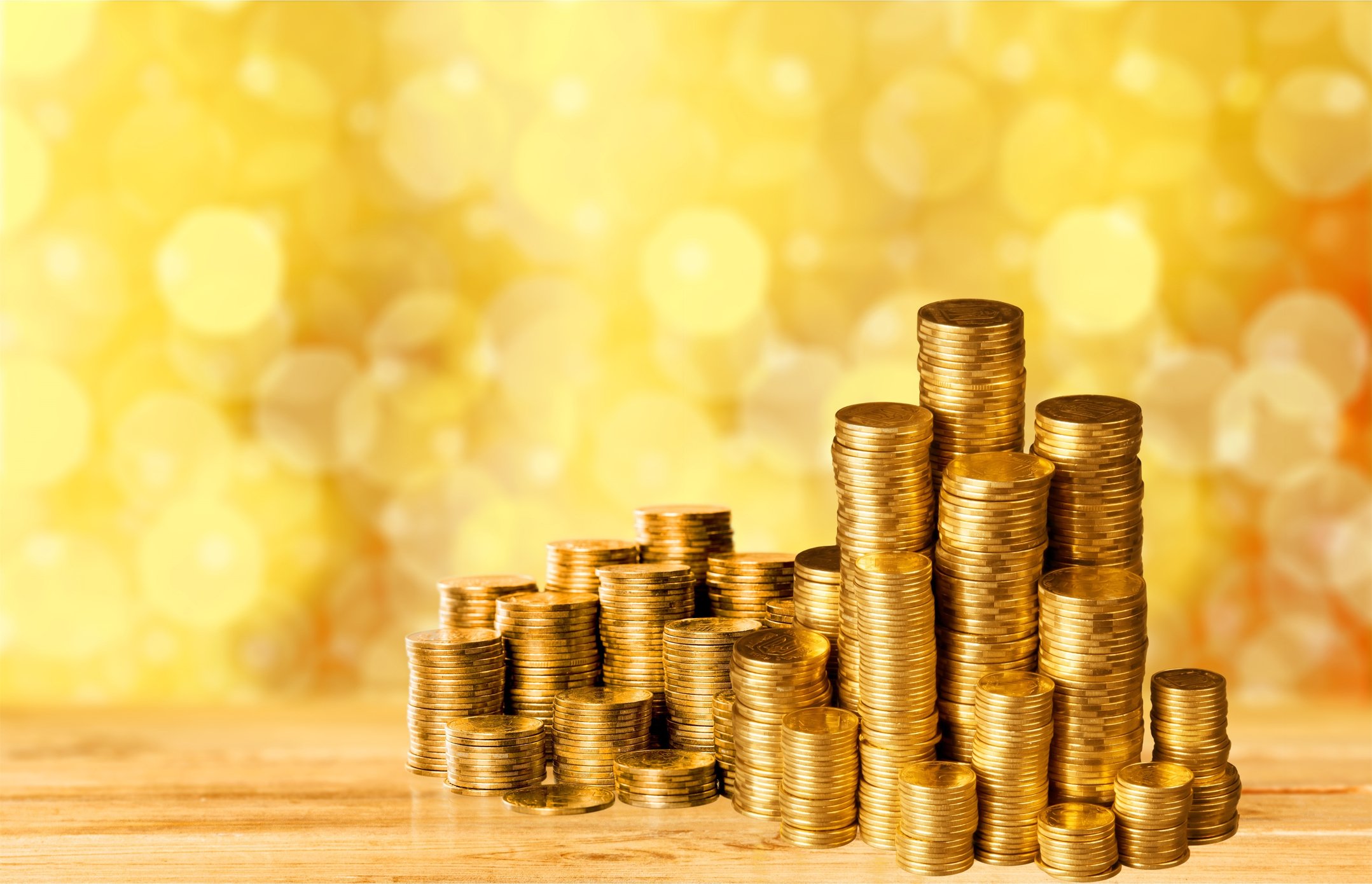 Many stacks of gold coins, some of which are very tall, in front of a glimmering gold background.