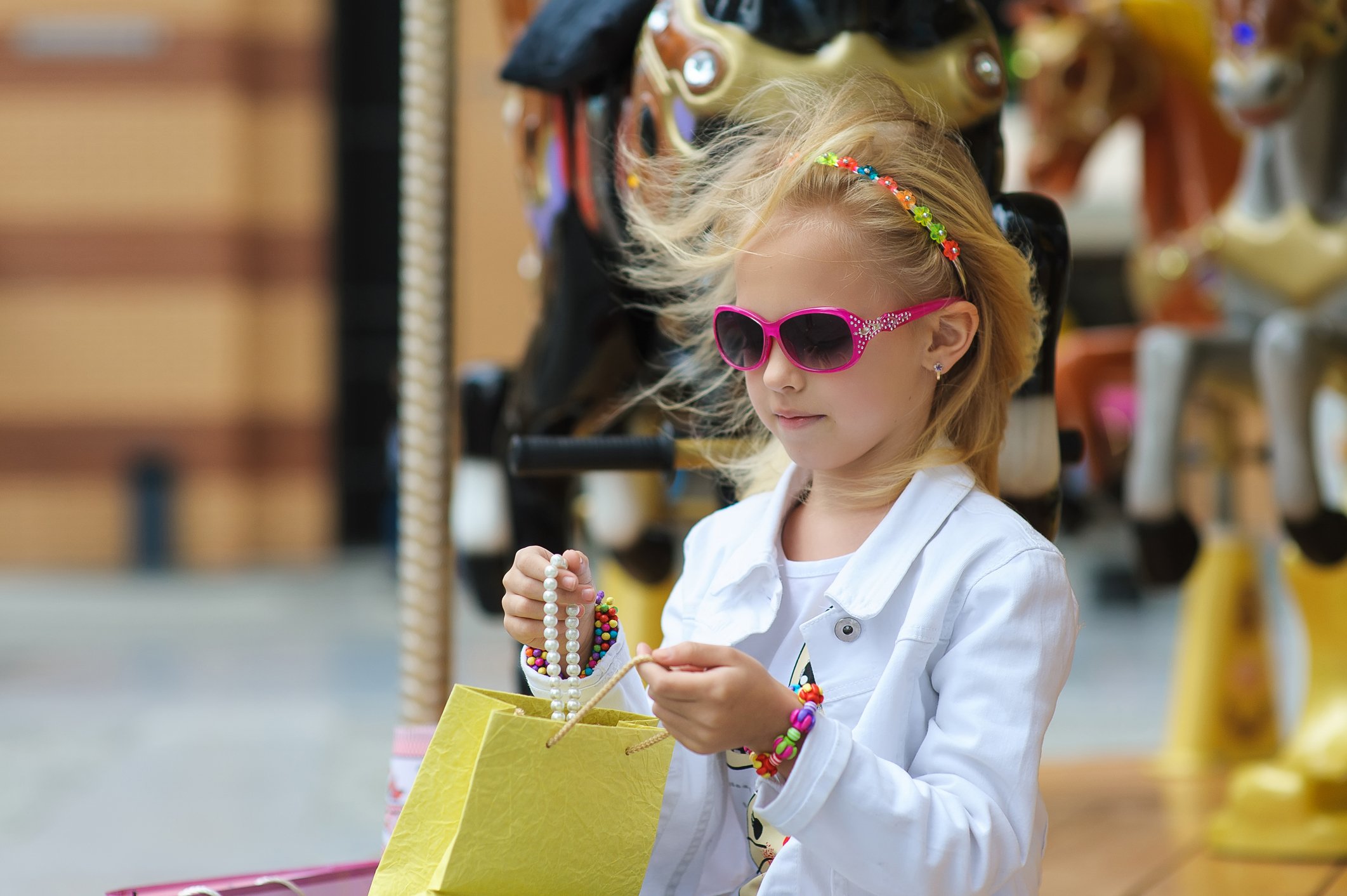 A young girl wearing pink sunglasses, bracelets, and a headband looks at jewelry she has in a yellow shopping bag