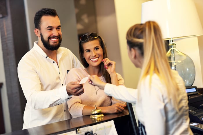 A woman behind a hotel desk hands a room key to a smiling couple.