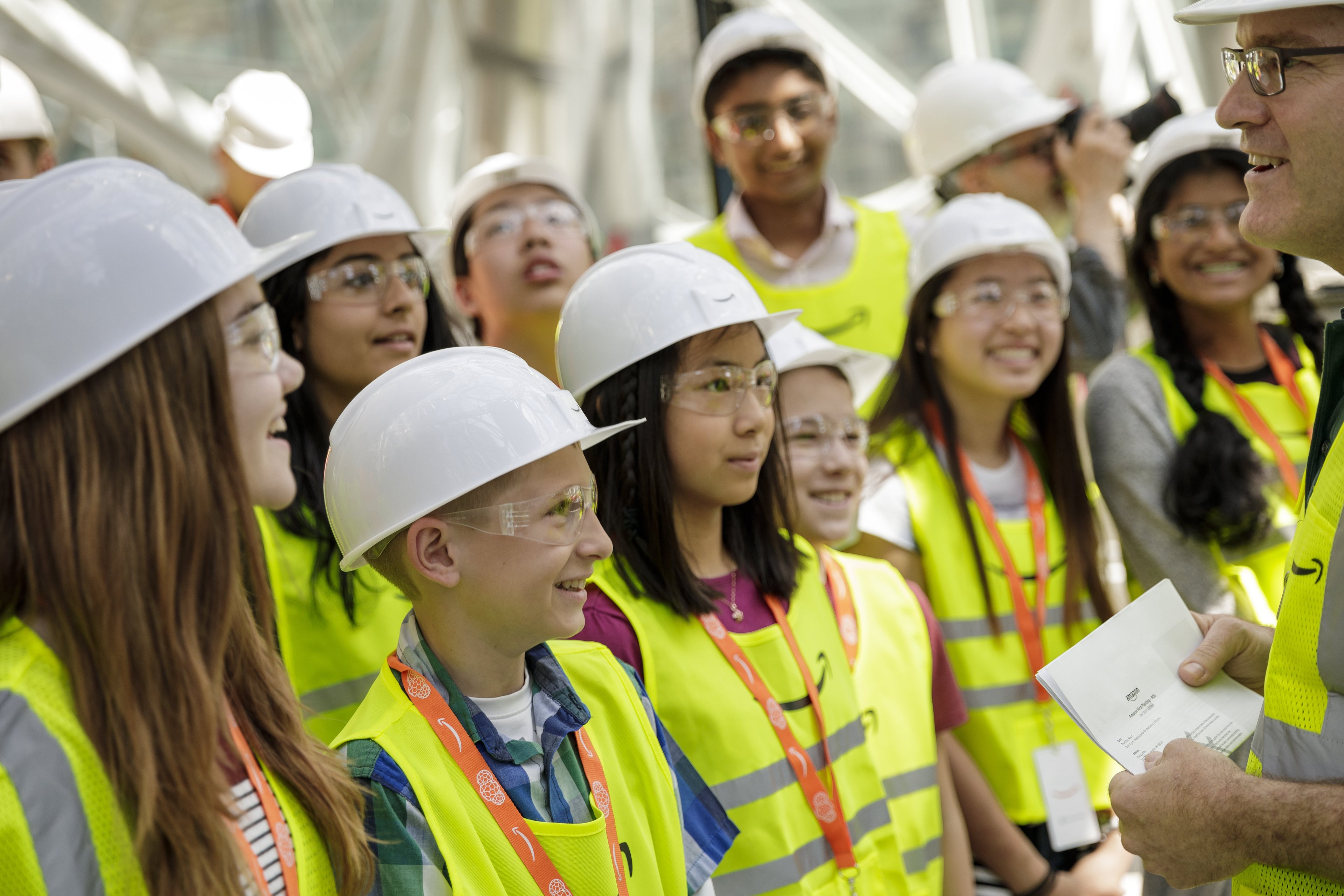 A group of Amazon employees in yellow vests and helmets faces a speaker.