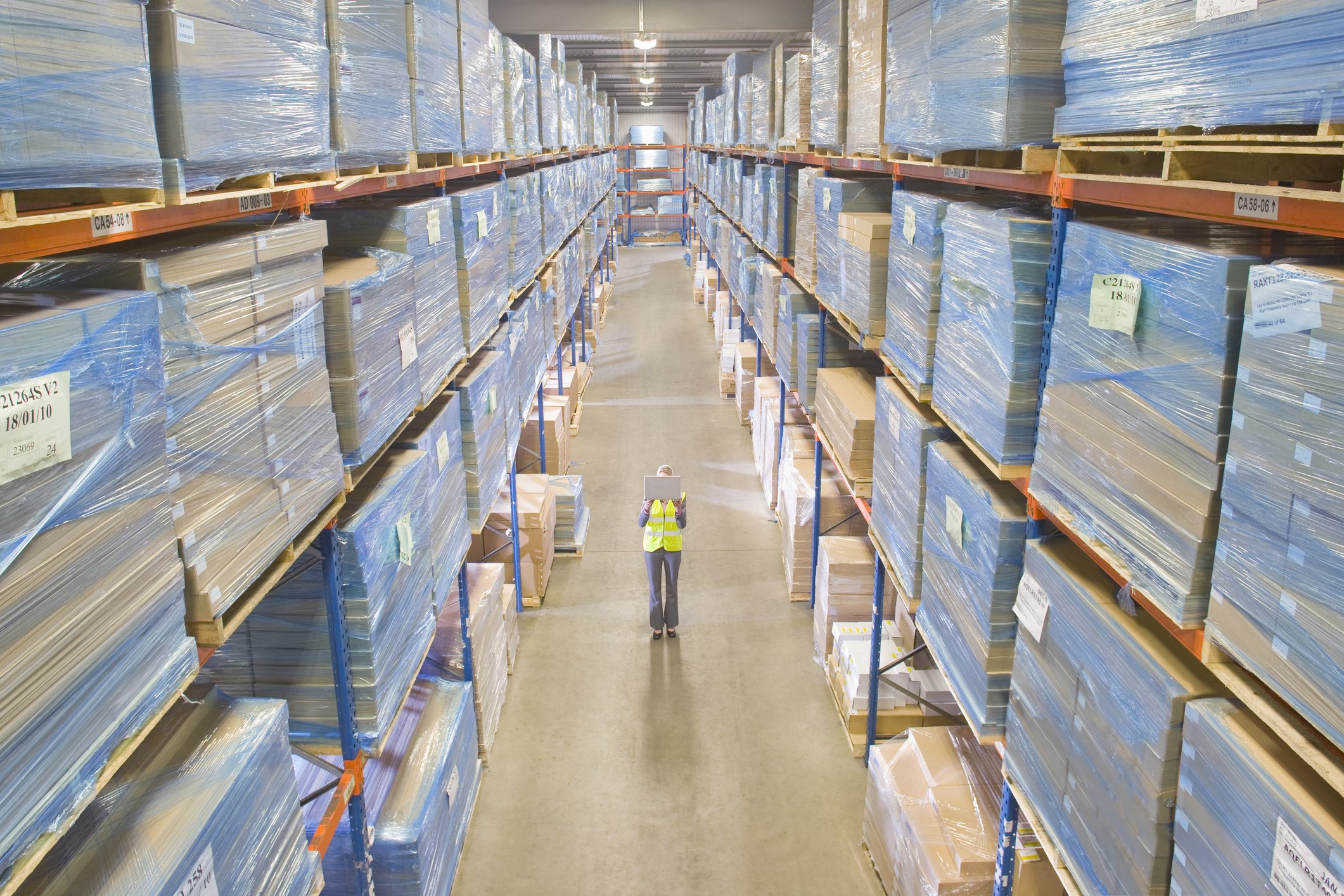 A warehouse worker holding a laptop in a warehouse that's filled with pallets of product.