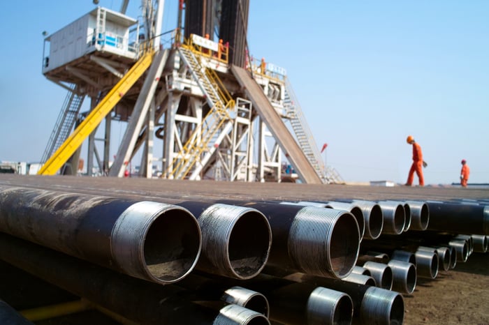 An oil rig with pipes in front of it and two workers walking in the back.