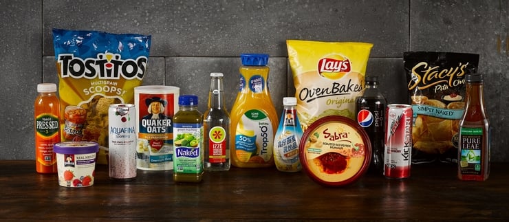 Assortment of 15 snack and beverage products on a table, including Tostitos, Lay's potato chips, an Izze beverage, Quaker Oats, Tropicana orange juice, Stacy's Pita Chips, and Sabra hummus.