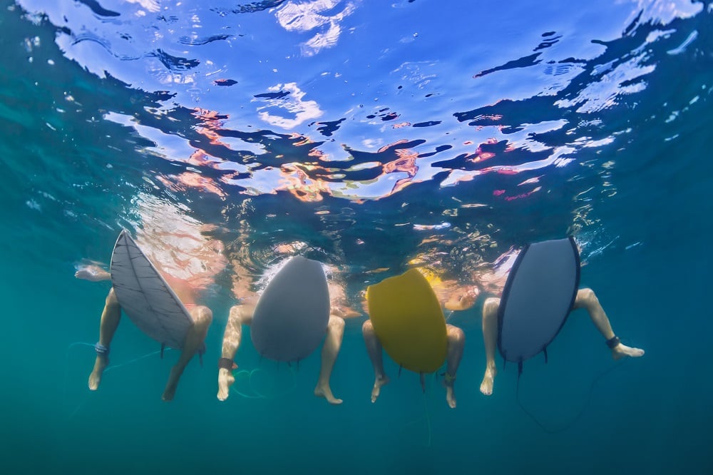 Surfers sitting on their boards with their feet in the water, as seen from below.
