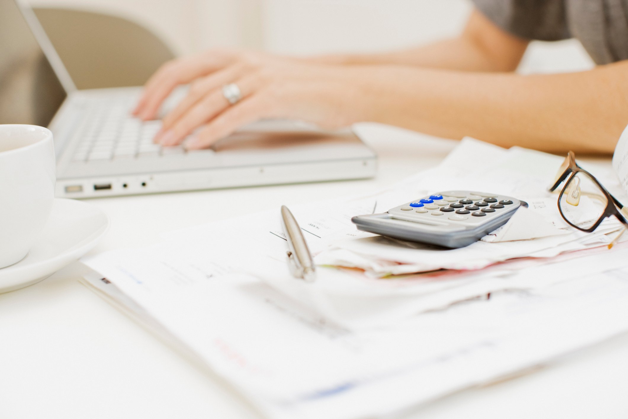 Person typing on a laptop with papers, a pen, glasses, and a calculator on hand