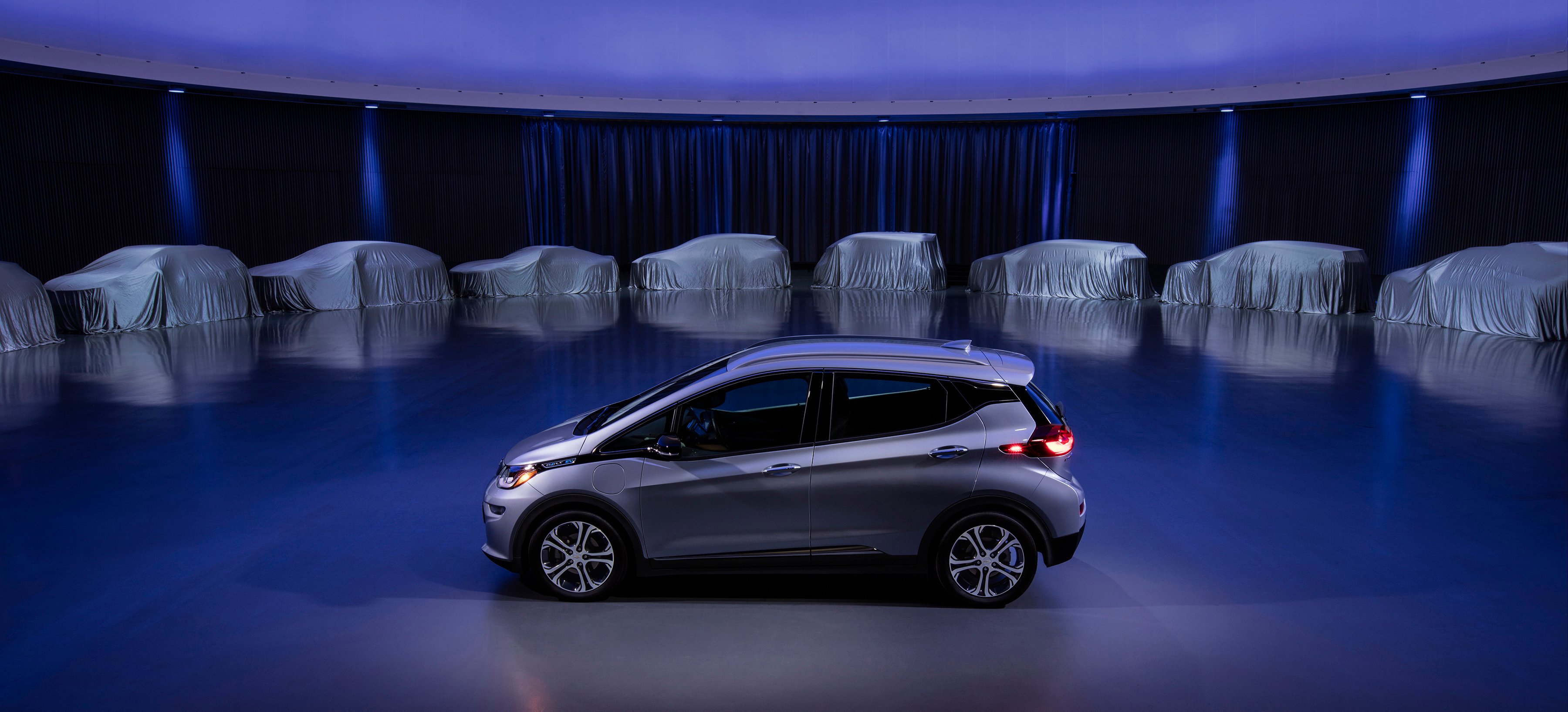 A silver Chevrolet Bolt EV is shown on a stage, with several covered vehicles in the background.