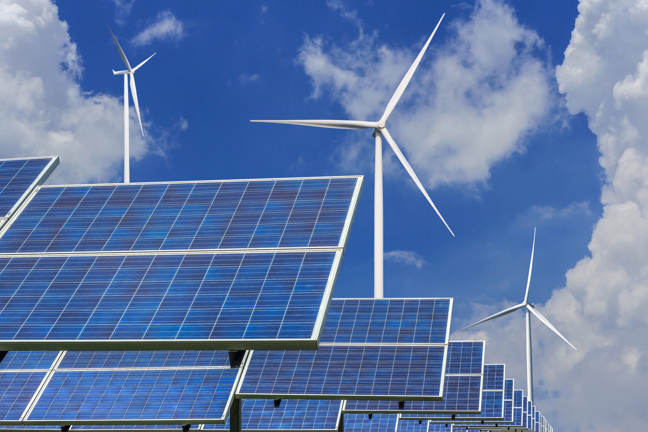 Several solar panels standing in front of some wind turbines.