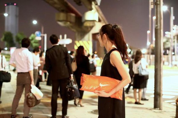 Woman in Japan on a busy sidewalk at night holding a bright orange Nike package.