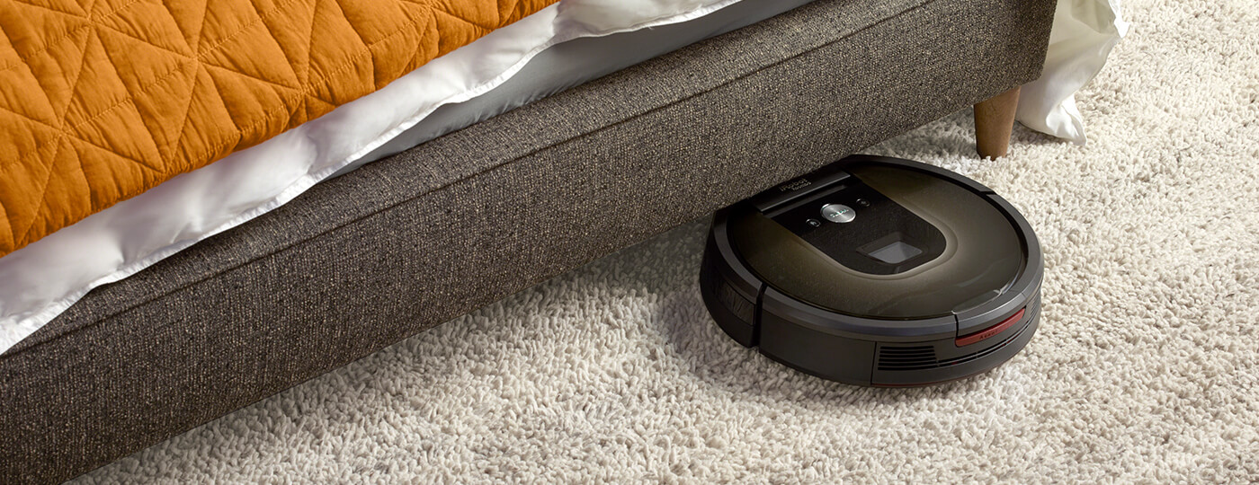A Roomba vacuum traveling on a rug and just about to go under a sofa.