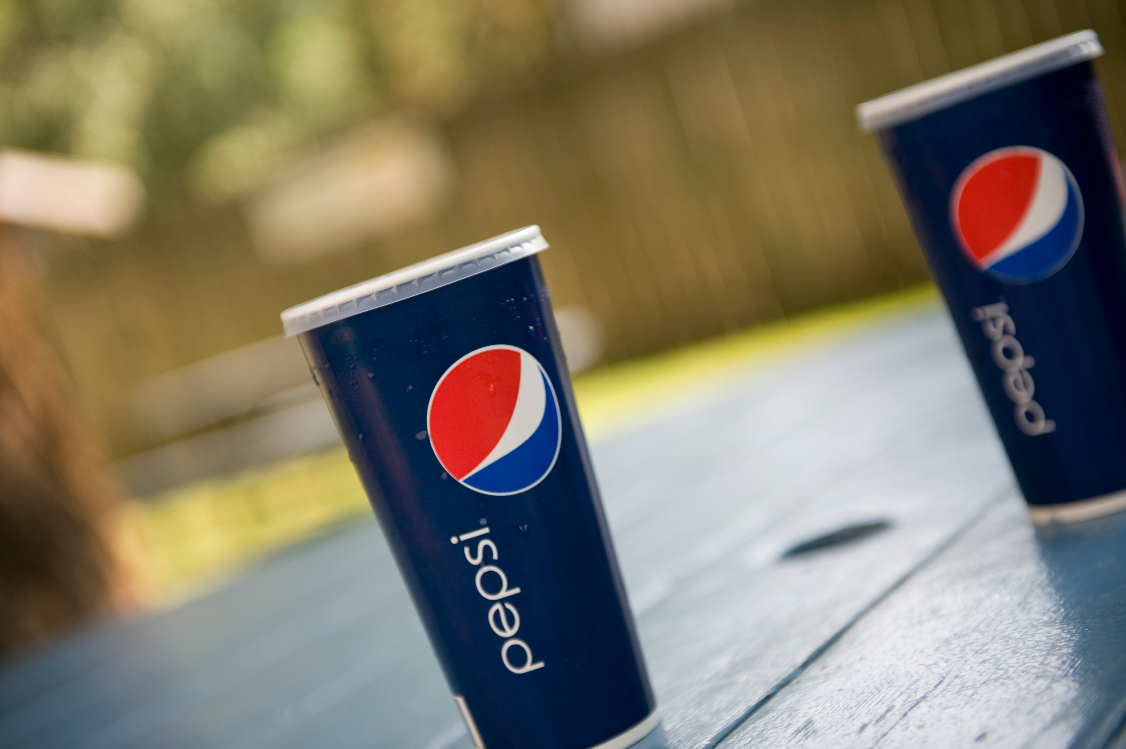 Two cups of Pepsi on a wood picnic table in a fenced-in backyard.