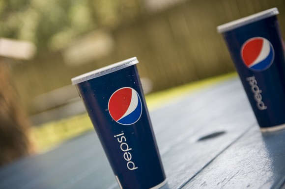 Two cups of Pepsi on a wood picnic table in a fenced-in backyard.