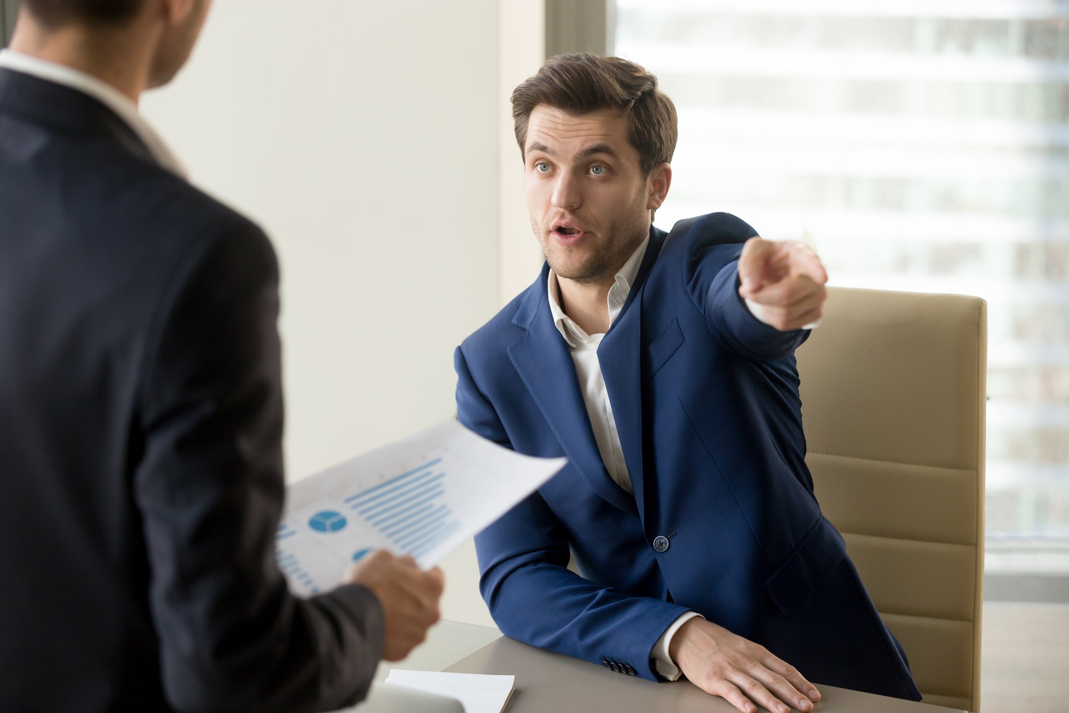 Man in suit pointing toward the door while meeting with another man holding a document