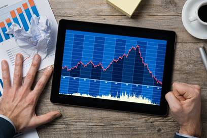 A person bangs his fist on a desk while looking at a tablet displaying a declining stock price chart.