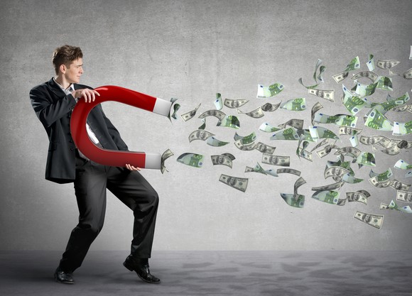 A man in a suit holding a large horseshoe magnet that is pulling dollar bills towards him.
