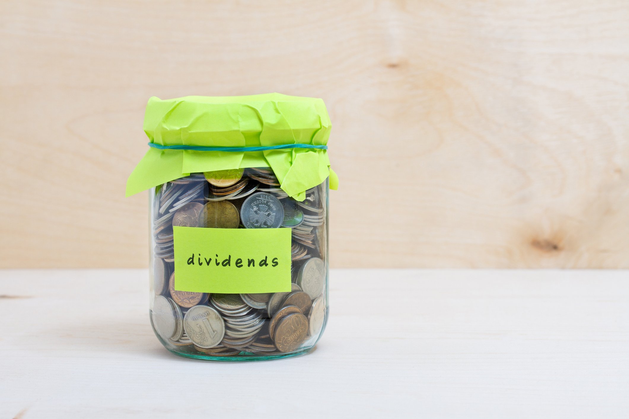 A jar filled with coins is closed with a rubber band around a piece of paper. The jar is labeled with the word "dividends."