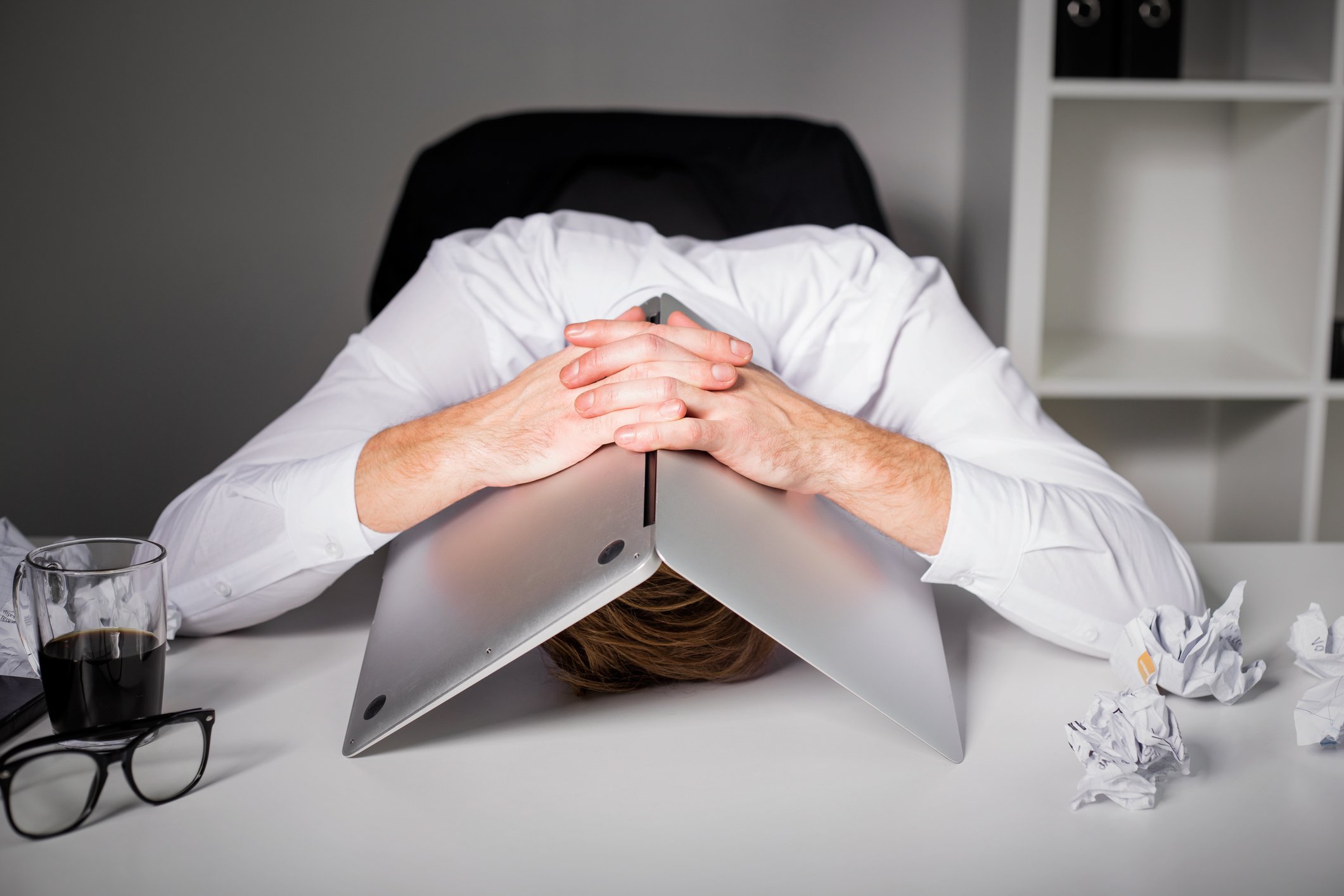 A worker with his head on his desk covered by his laptop.