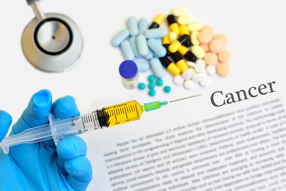 Cancer pills on top of document with end of stethoscope and hand holding syringe