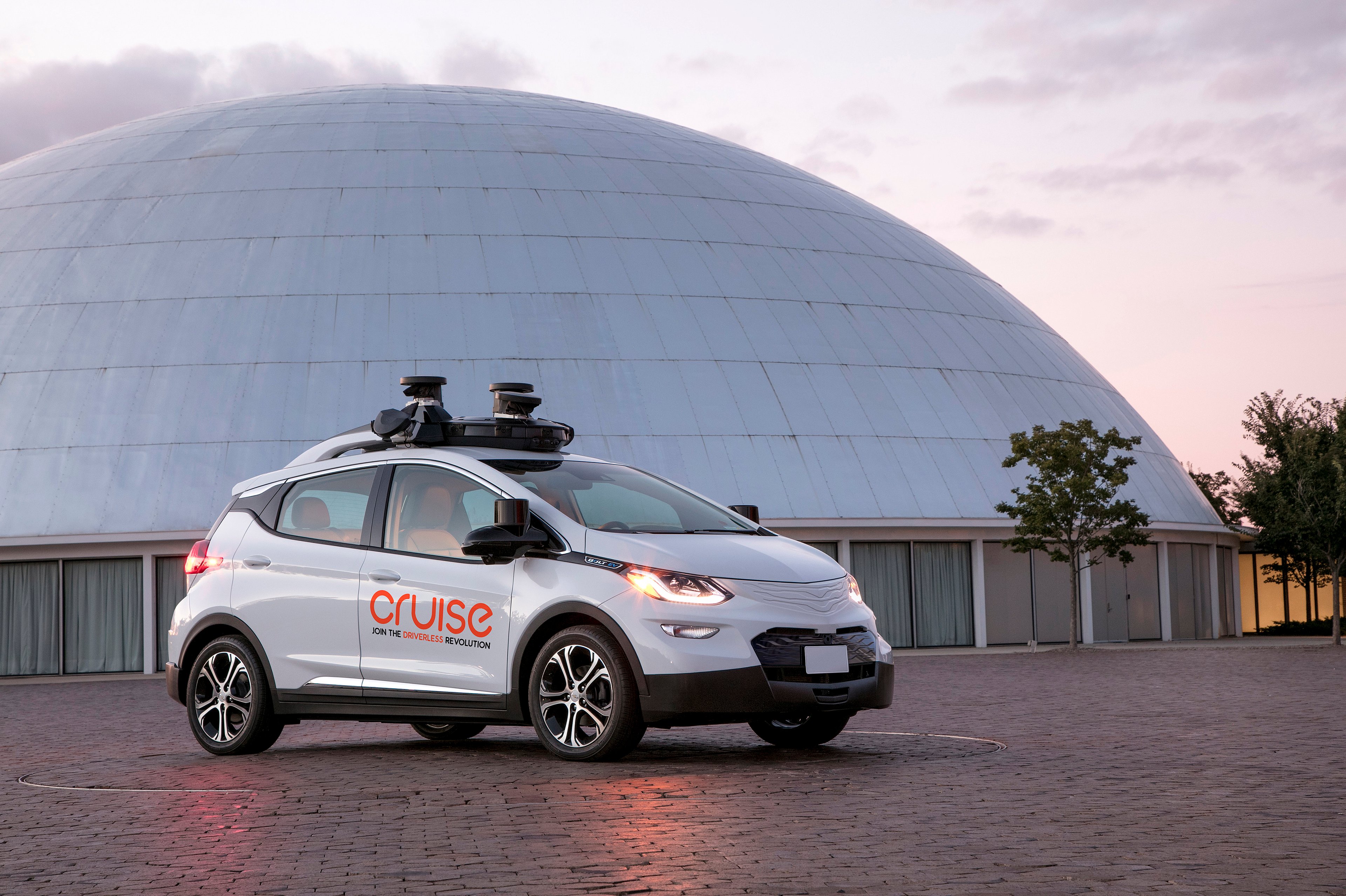 A white Chevrolet Bolt EV with Cruise logos and visible self-driving sensor hardware is parked next to GM's historic Design Dome in Warren, Michigan.