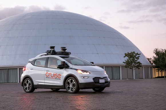 A white Chevrolet Bolt EV with Cruise Automation logos and visible self-driving sensors is parked next to a dome.