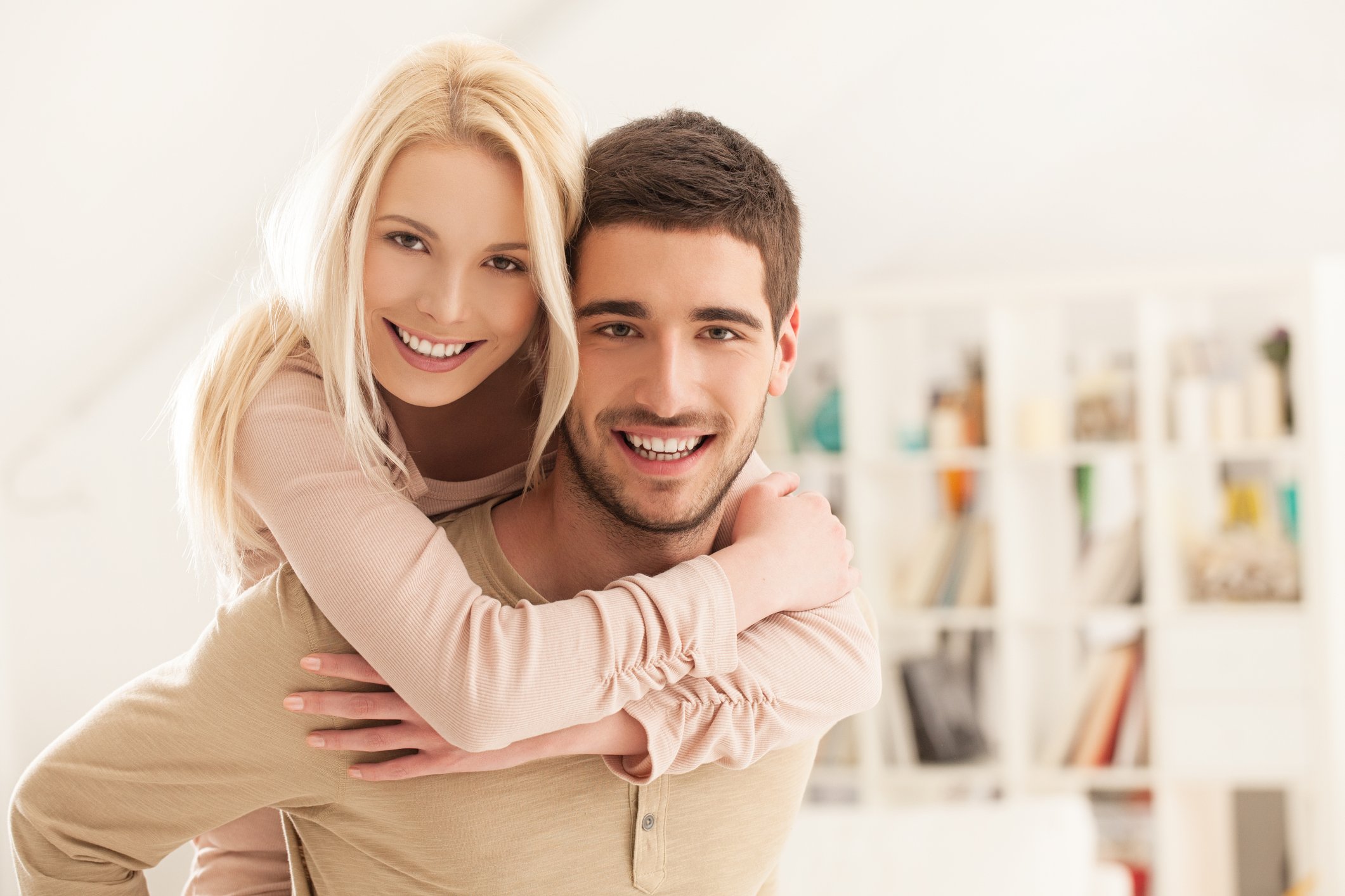 A young couple smiles at the camera, with the woman's arms wrapped around the man's as she stands behind him. In the background is a bookcase full of books.