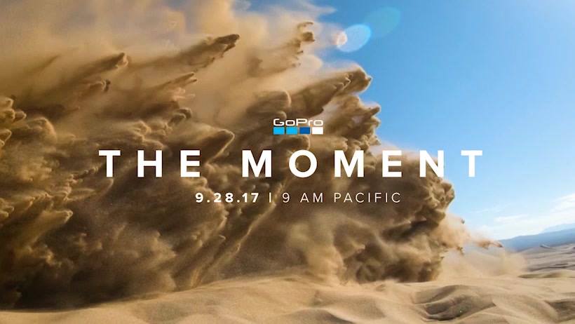 Flying sand kicked up by a motorcycle riding in the desert with a blue sky background and the words The Moment in the foreground with the date and time underneath and a GoPro logo.