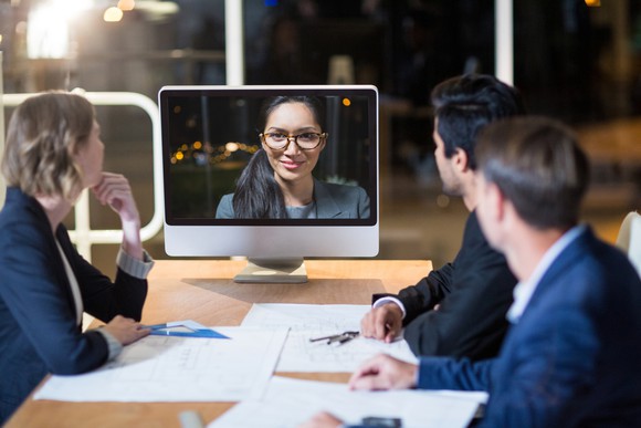 In a teleconference, three people in business attire are gathered around a table, with papers laid out on the table between them, as they look at a person on a monitor.
