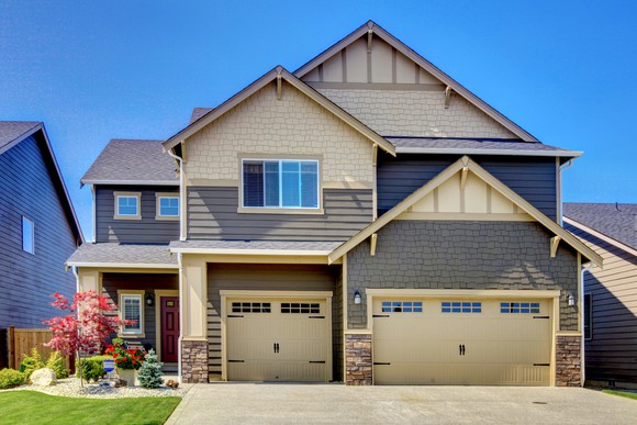 A two-story house with a double garage, green lawn, and large driveway against a blue sky.