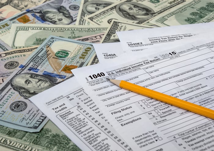 Tax forms and a pencil on top of a shallow pile of $100 bills.