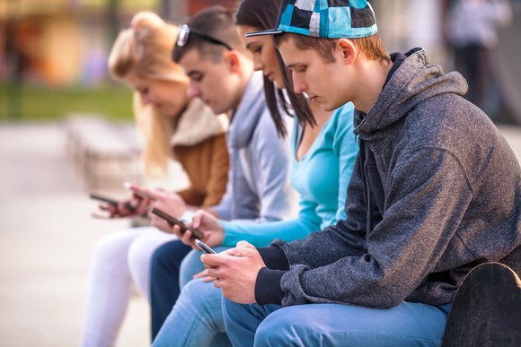 Teenagers sitting on a bench all using their smartphones.