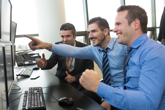 3 men in shirts and ties in an office setting smile as one points to a computer monitor.