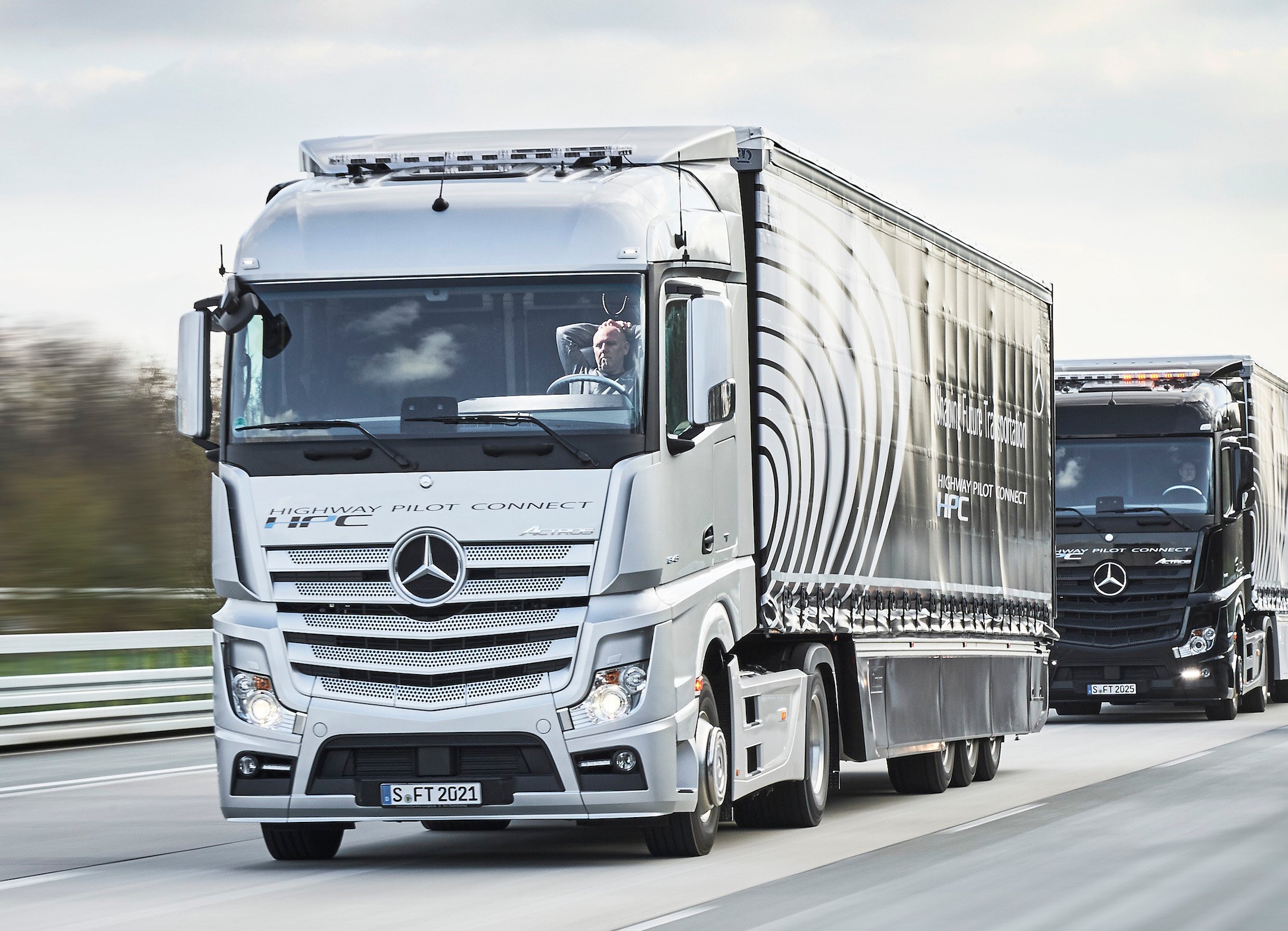 Two Mercedes-Benz tractor-trailer trucks on a highway. The lead truck's human driver has his hands behind his head to show that the trucks are driving themselves.