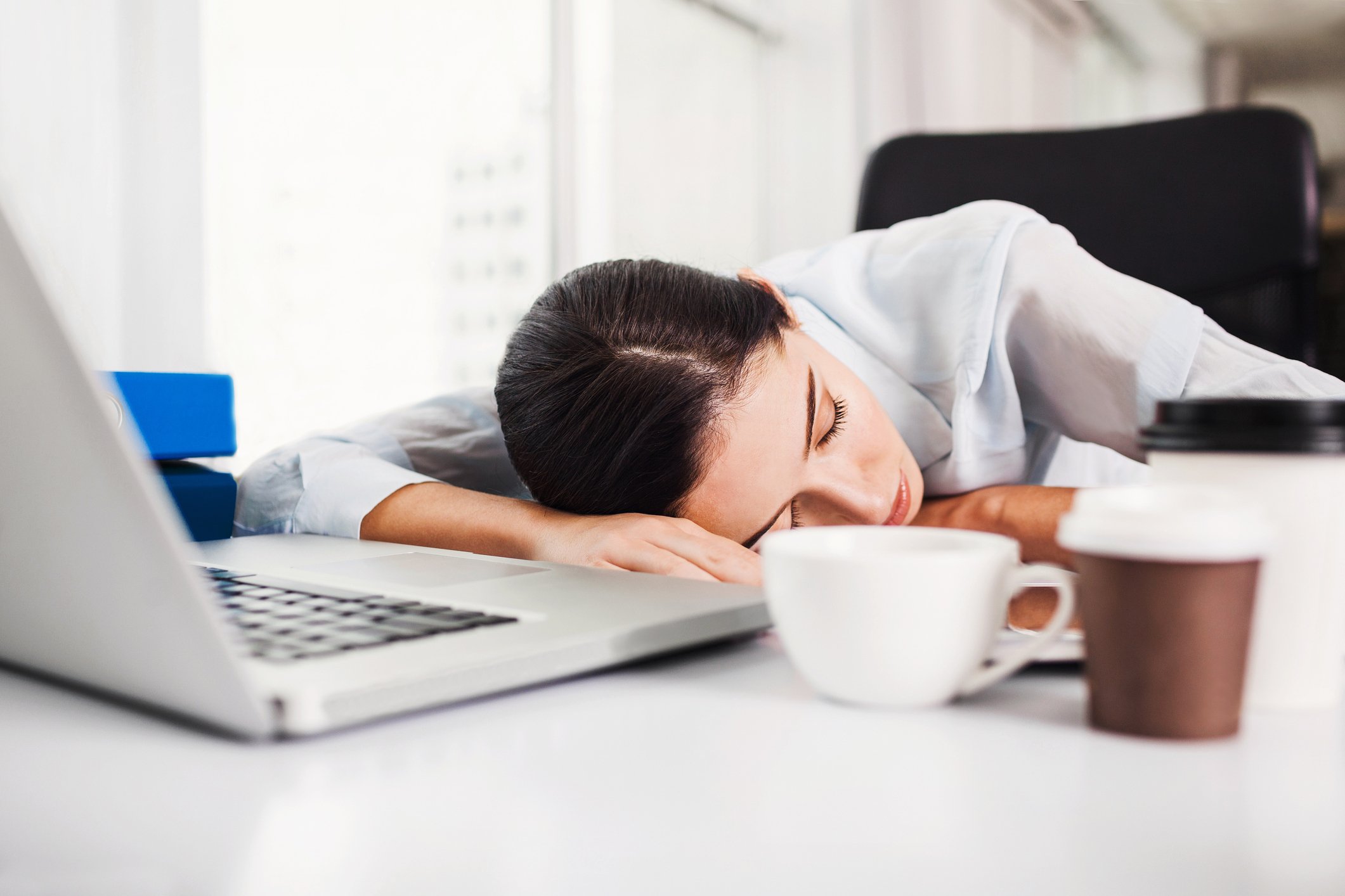 A woman sleeps at a desk with a computer and coffee cups on it.