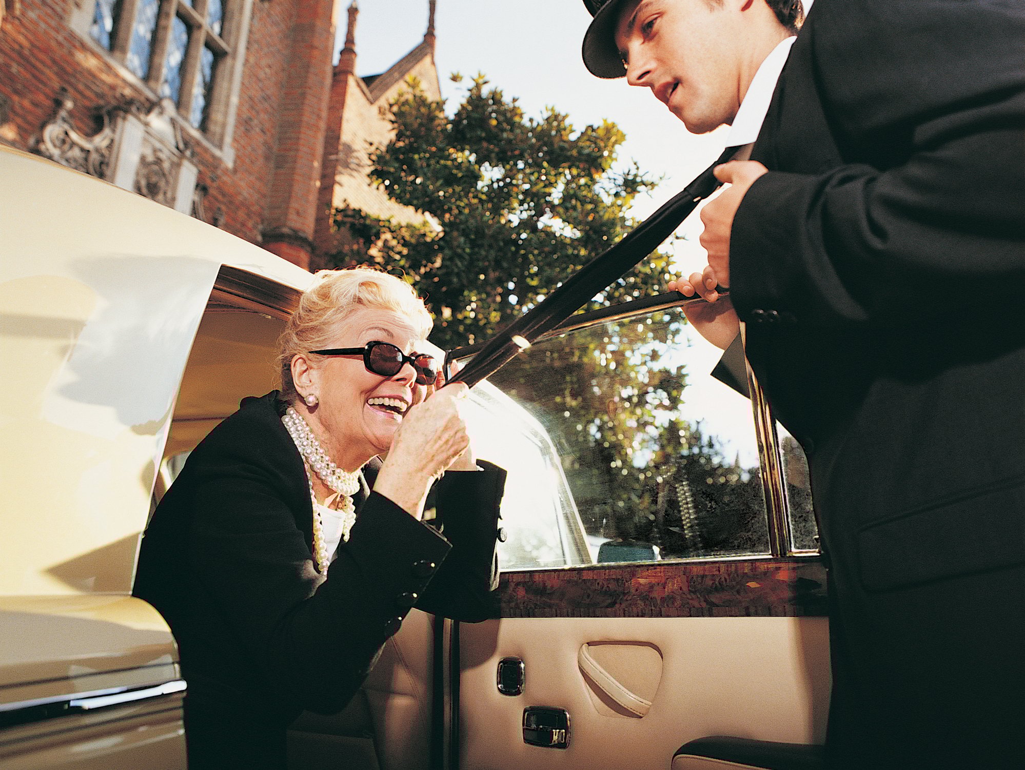 Driver opening car door for older woman in black jacket and pearls