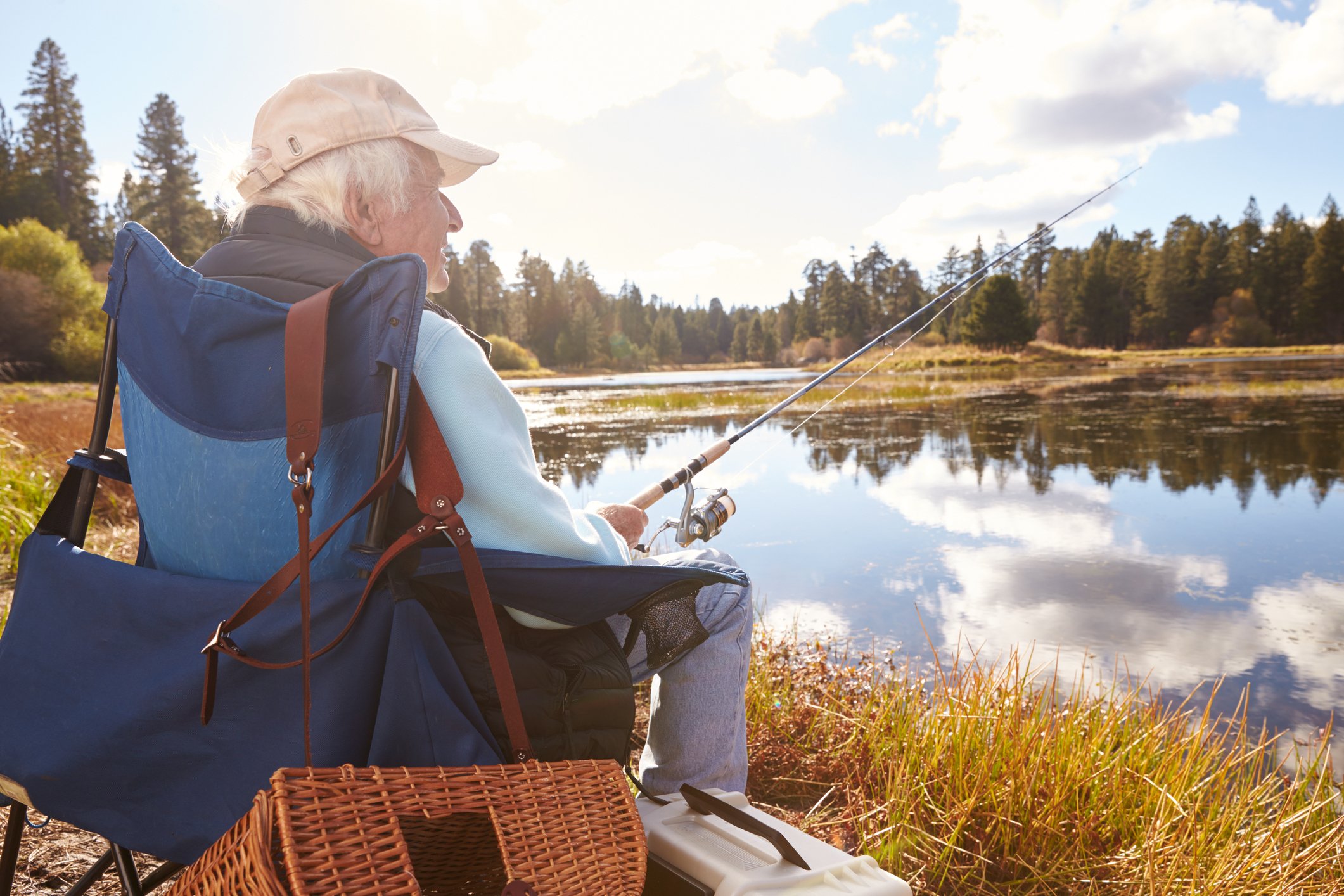 An old man wearing a ball cap ad sitting in a chair fishes from a pond.