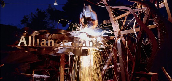 Welder working on a metal apparatus shown with an Alliance Bank logo in front of it.