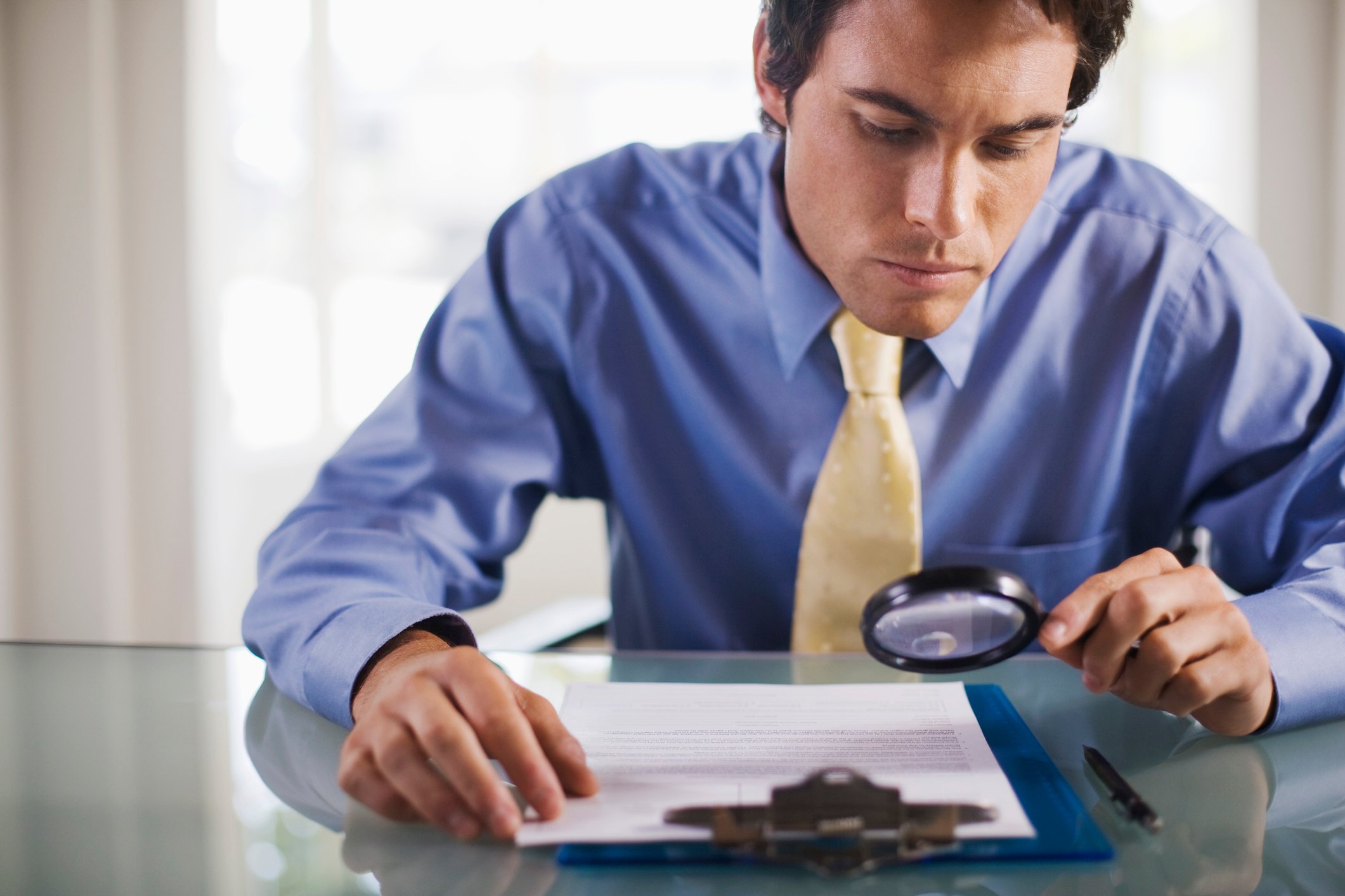 A man in a button-down and tie studies a document with a magnifying glass.