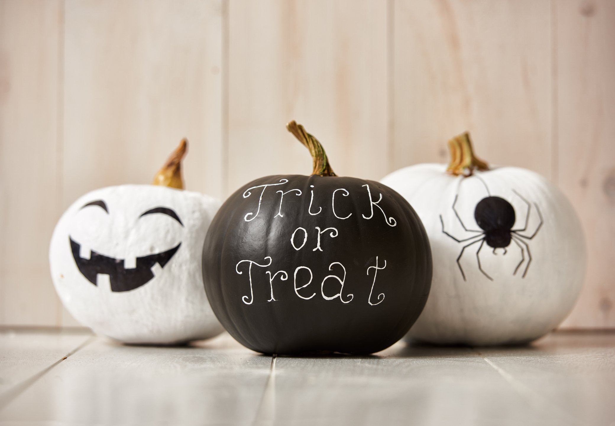 Three pumpkins are decorated for Halloween. The white pumpkin on the left is a smiling jack o'lantern, the black pumpkin in the center reads "trick or treat," and the white pumpkin to the right has a drawing of a spider.