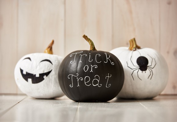 Three pumpkins are decorated for Halloween. The white pumpkin on the left is a smiling jack o'lantern, the black pumpkin in the center reads "trick or treat," and the white pumpkin to the right has a drawing of a spider.