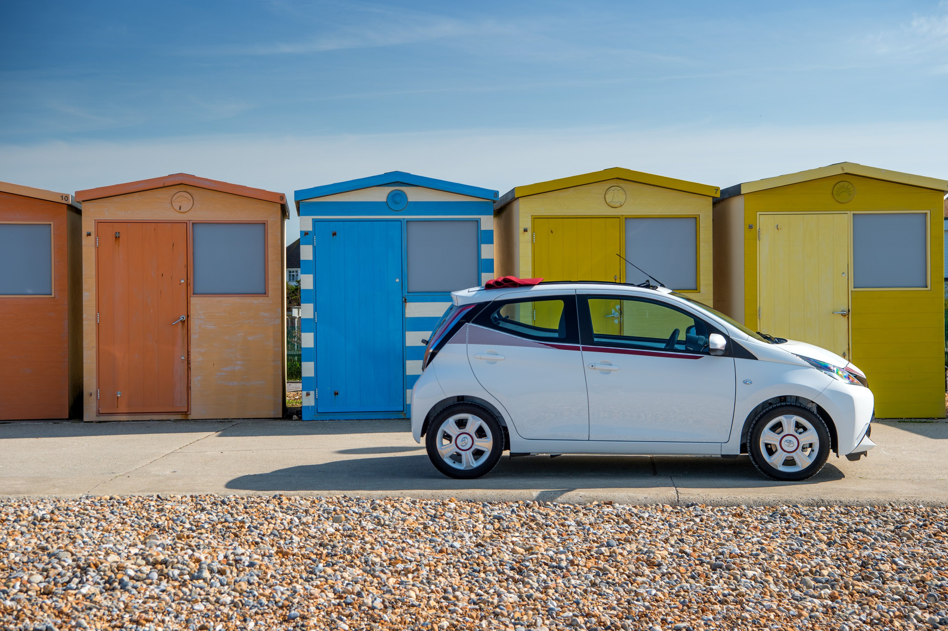 A white Toyota Aygo x-claim exterior pictured in front of colorful sheds on a beach