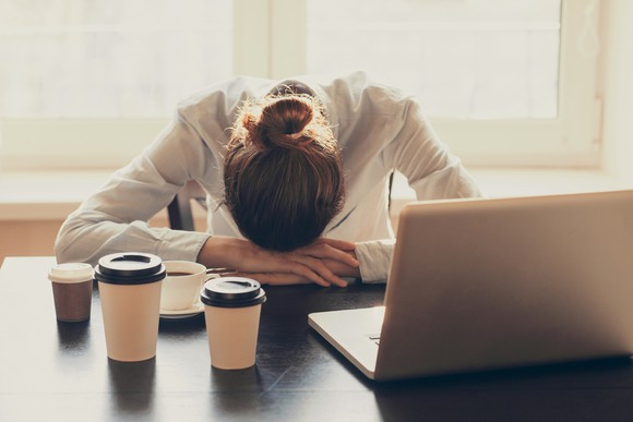 A woman has her head down on a desk with cups of coffee and a laptop in front of her.